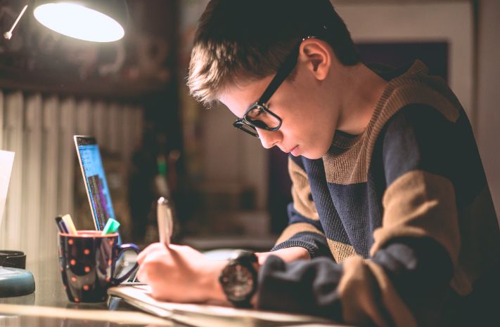 High school-aged boy wearing glasses studying in his bedroom with a laptop, taking notes in a spiral notebook.