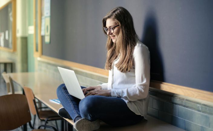 Female high school student sitting on table near chalkboard using a white laptop.