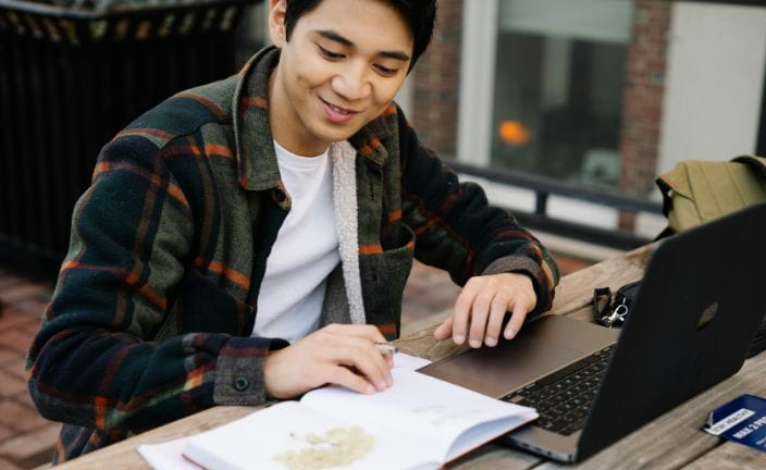 Man in plaid shirt studying with notebook and laptop in office.