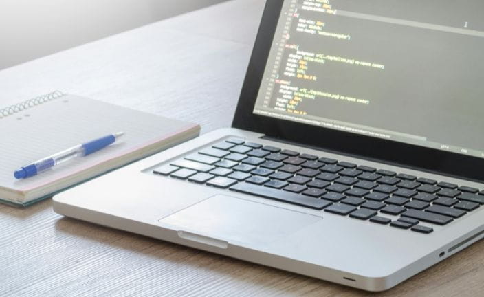 Laptop on desk displaying code next to notebook with blue pen.