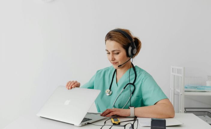 Woman in scrubs wearing headset opening laptop.